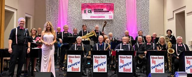 The Robson Ranch Big Swing'n Band posed on stage with their music stands, dressed in black with pink accents, in front of a silver backdrop, pink drapes, and a Women Rock banner
